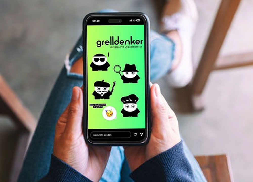 Das Bild zeigt eine Person, die ein Smartphone in der Hand hält. Auf dem Bildschirm des Telefons sind verschiedene Charaktere in einem Cartoon-Stil zu sehen, die scheinbar Teil eines digitalen Stickersets sind. Jeder Charakter trägt einen schwarzen Hut und eine Brille, und einer hält sogar eine Lupe. Sie repräsentieren wohl die Marke oder das Team von "grelldenker", was als Text über den Charakteren auf dem Bildschirm steht, zusammen mit dem Zusatz "die kreative Digitalagentur". Das Bild fängt einen alltäglichen Moment ein, möglicherweise um die Verwendung der Sticker in einer Messaging-App zu zeigen.